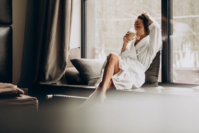 The image shows a woman sitting on a window seat in a relaxed pose, wearing a white bathrobe. 